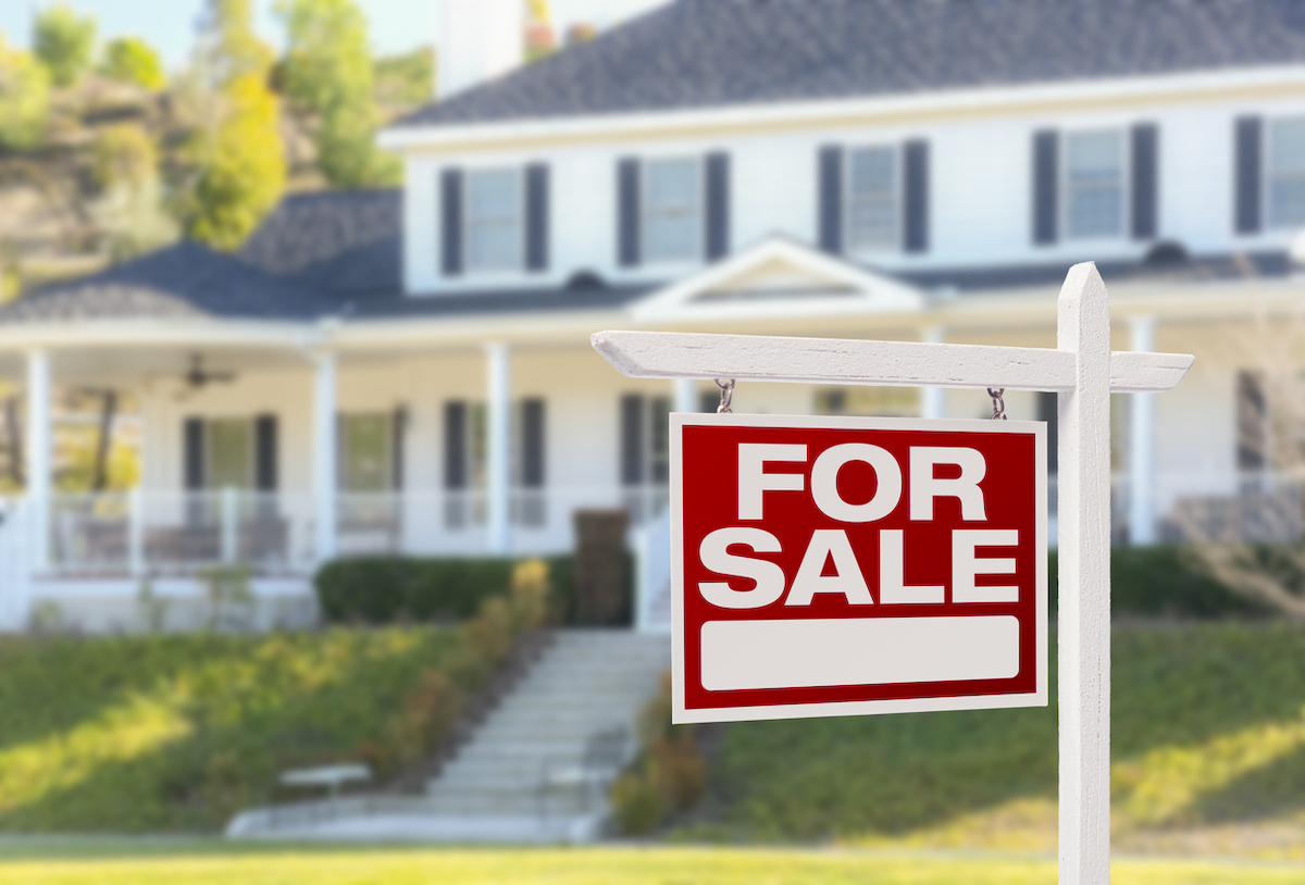 A for-sale sign in the foreground with a big white house in the background