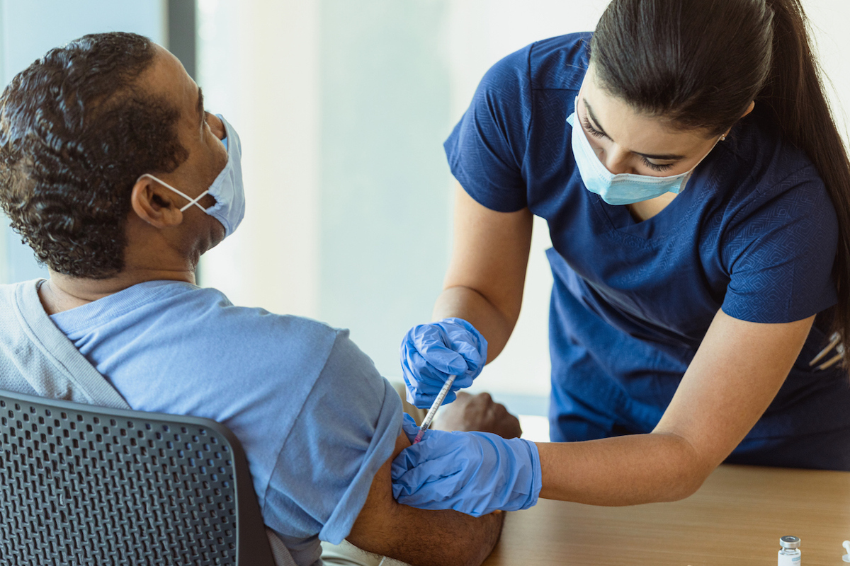 A man receives his first dose of the COVID-19 vaccine from a female healthcare professional. They are both wearing protective masks.