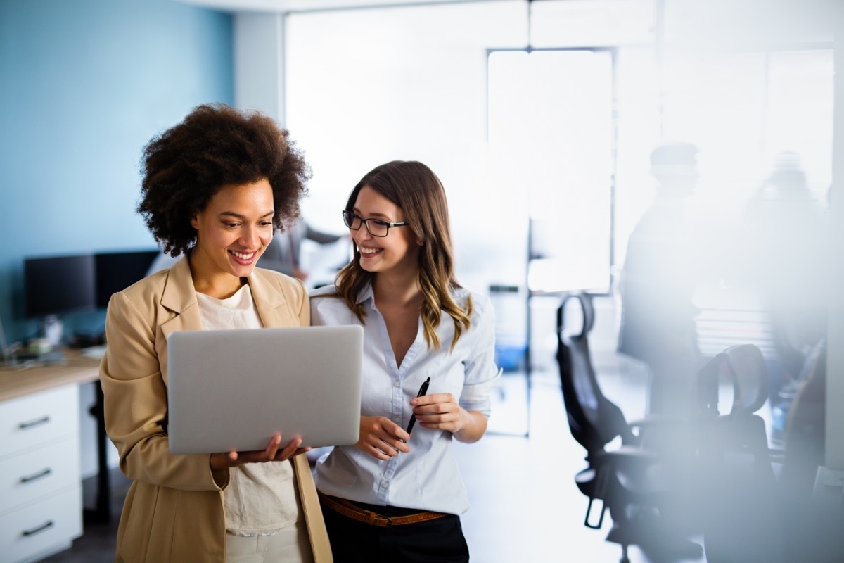 two female coworkers at the office, smiling and looking at a laptop while standing