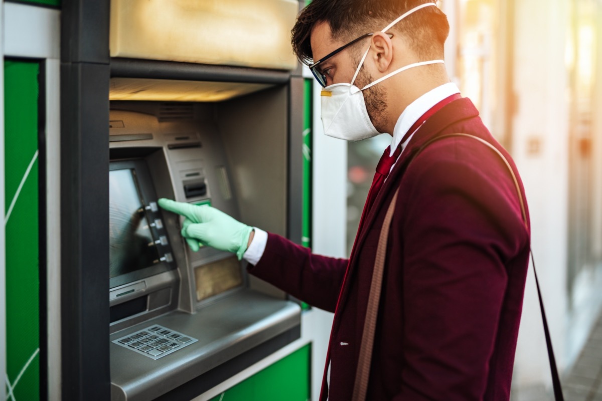 Elegant young man with protective mask standing on city street and using ATM machin with protective gloves on hands. Virus pandemic prevention and healthcare concept.