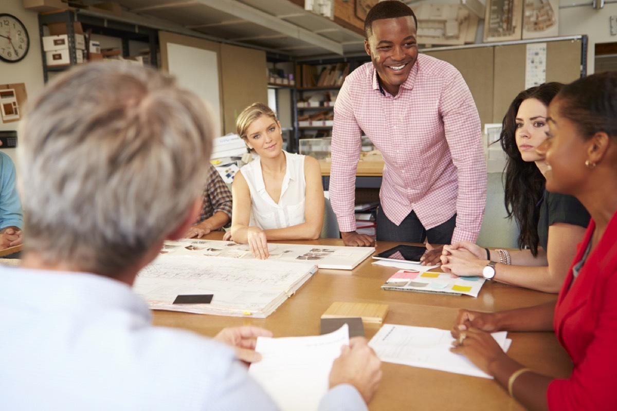 man standing up at desk with female employees seated, office etiquette