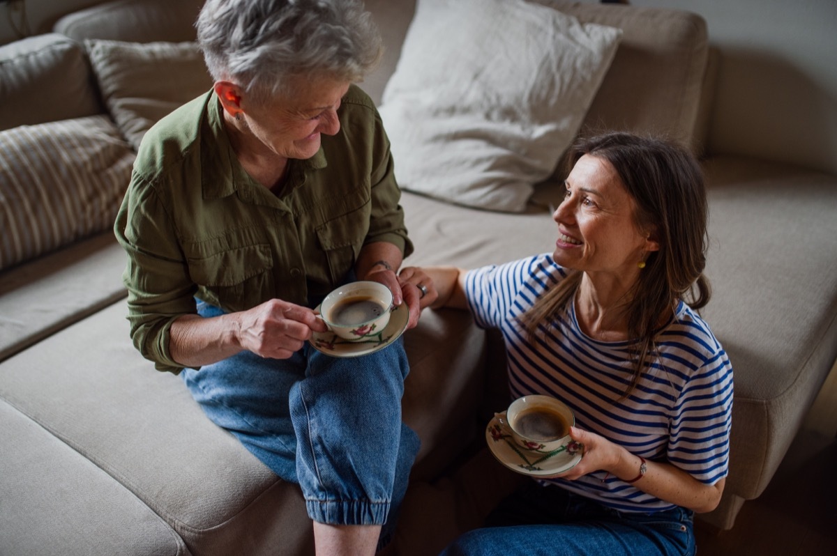 Happy senior mother drinking coffee with adult daughter indoors at home, talking.