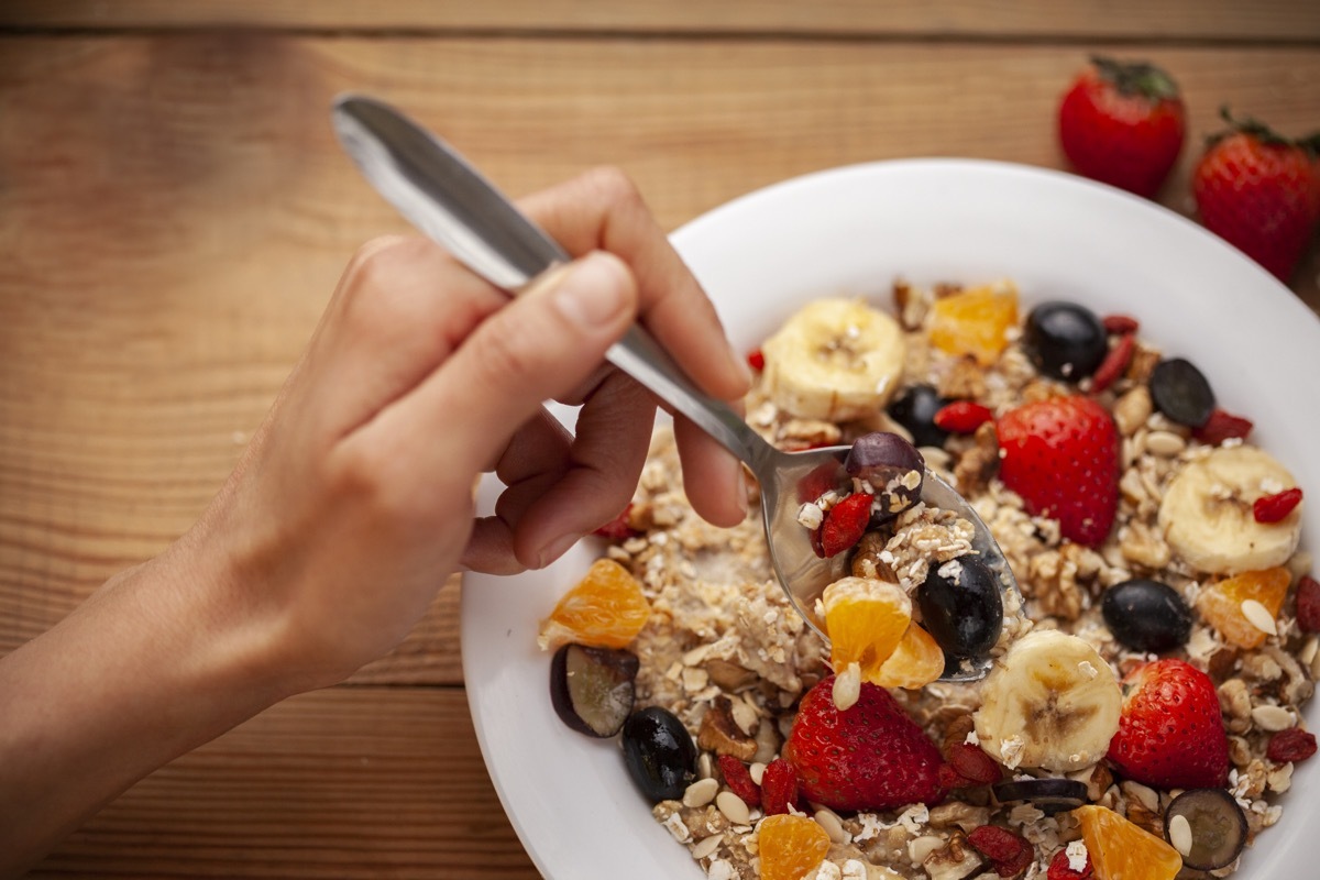 Top view close up of delicious, healthy homemade oats breakfast garnished with variation of mixed fruits toppings served in a white plate on wooden dining table and a hand picking a spoon full of it to eat.