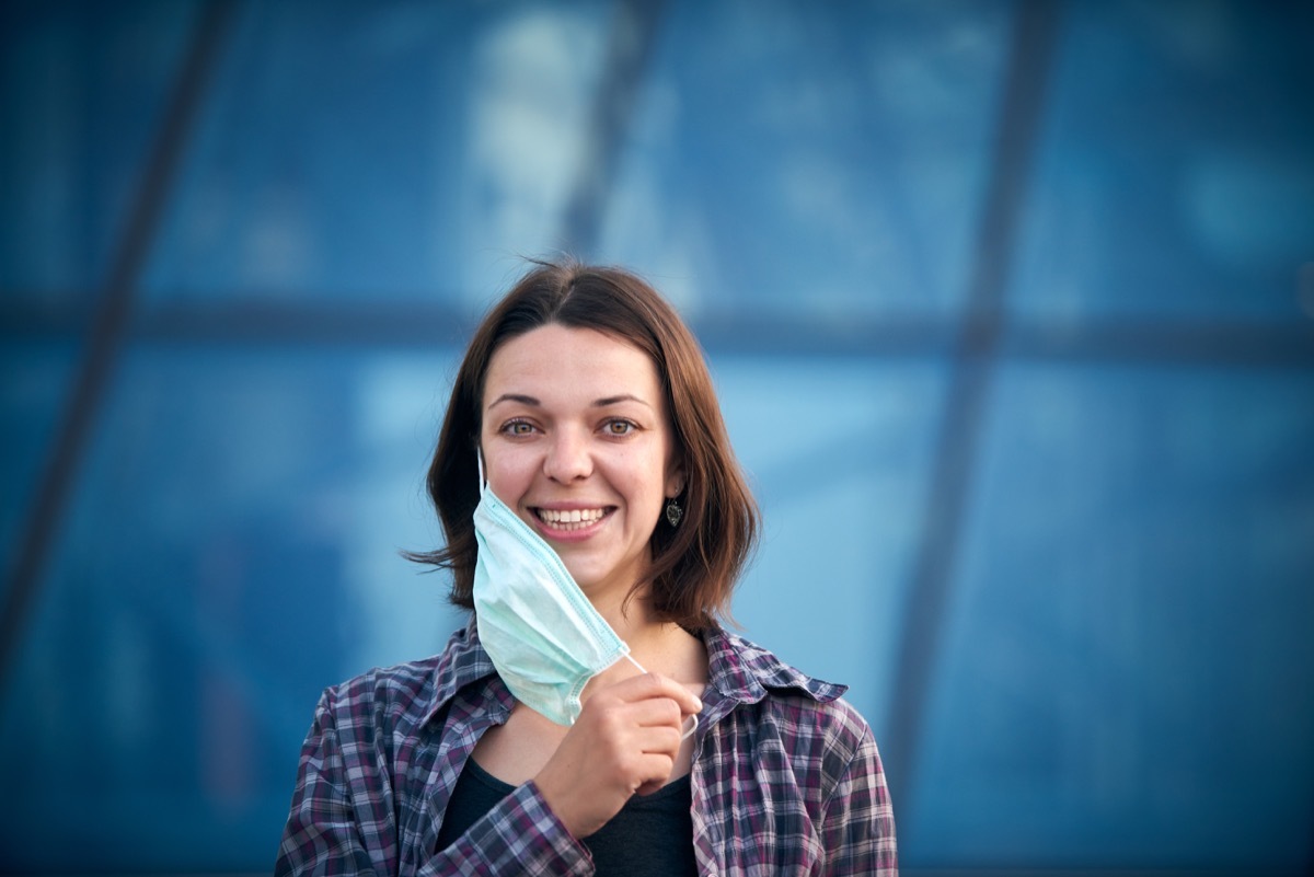 Happy woman takes off protective medical mask outdoors against modern city background. Pandemic Covid-19 is over.