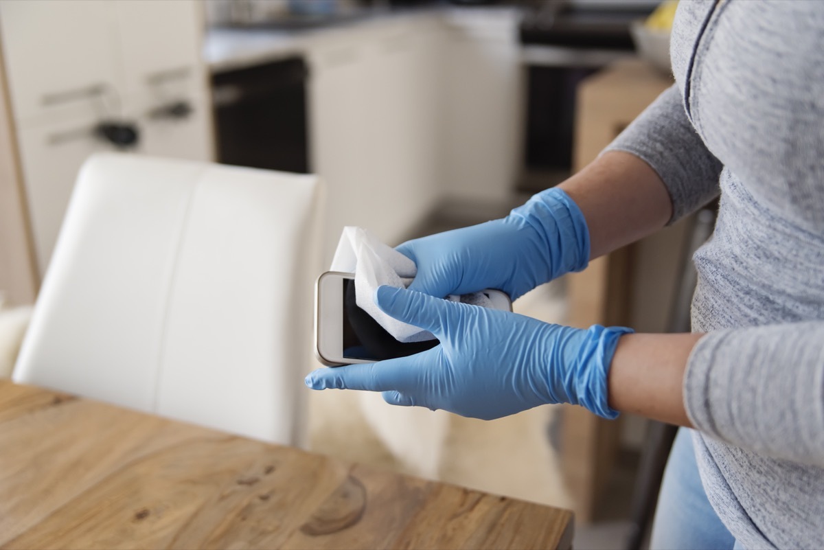 Female hands with blue glove wiping mobile phone with disinfectant wipe. Horizontal indoors close-up with copy space.