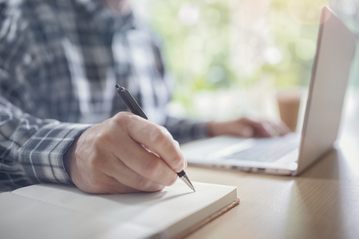 man writing in pad next to laptop