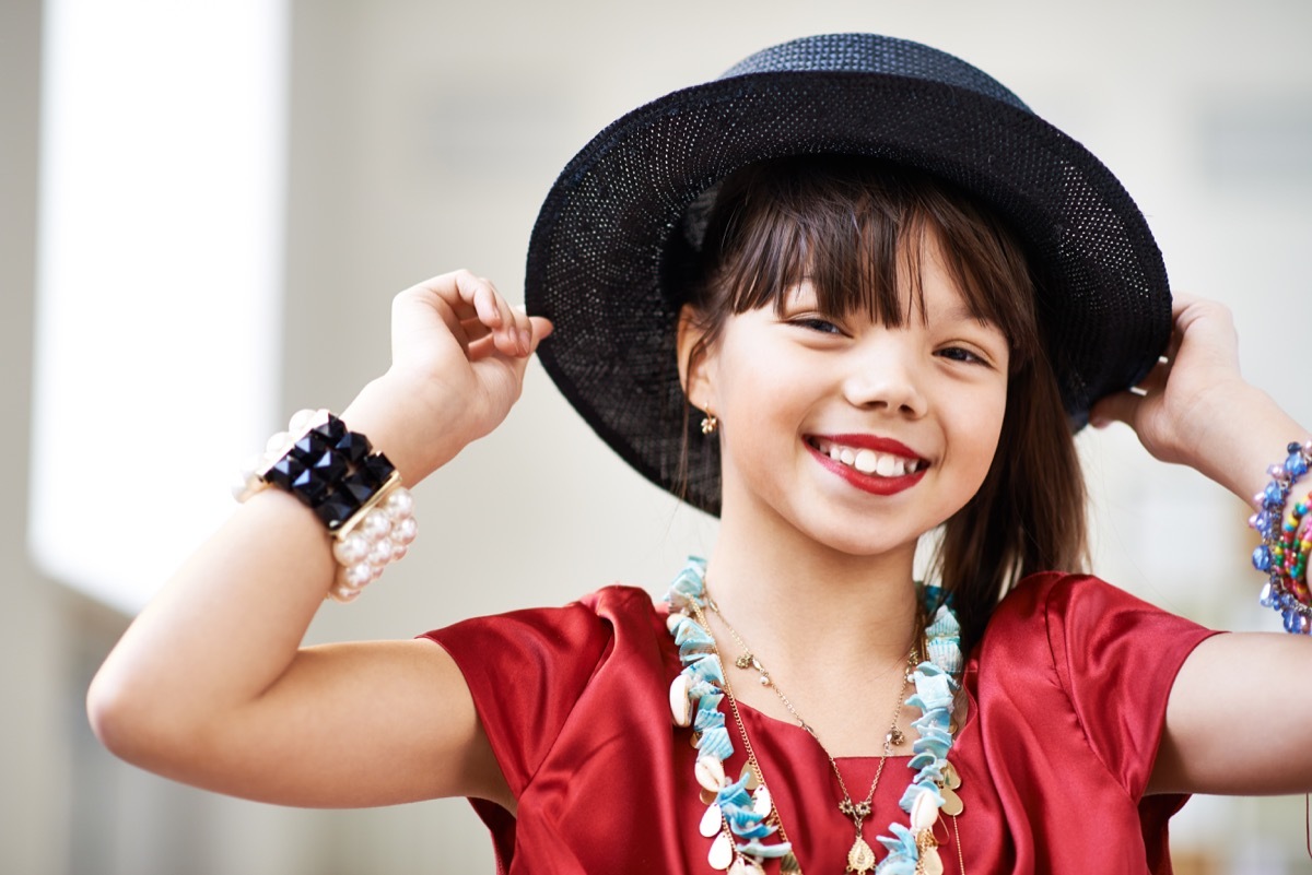 Charming girl with red lipstick wearing adult dress with hat and accessories looking at camera