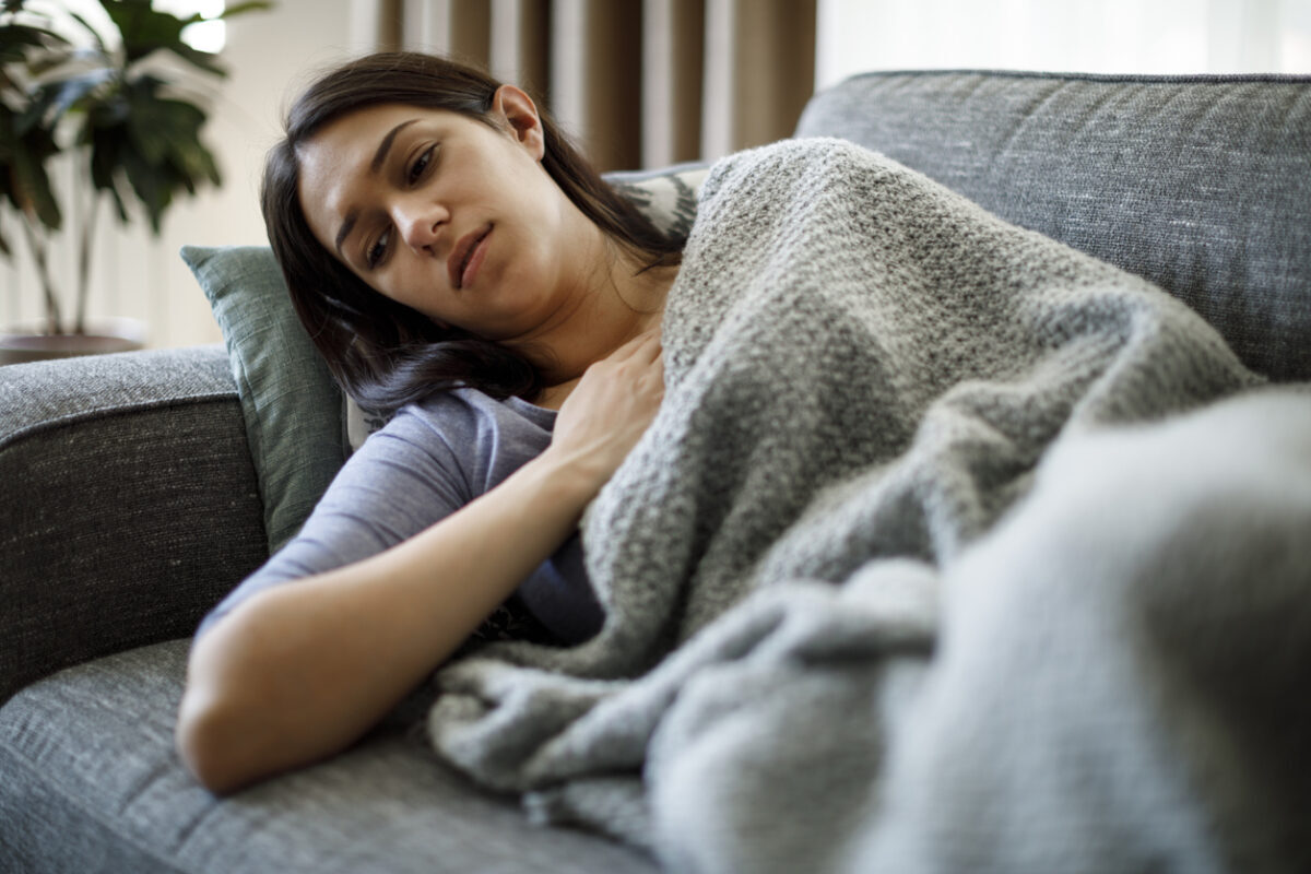 A young woman lies on the couch while covered by a blanket and a tired expression on her face