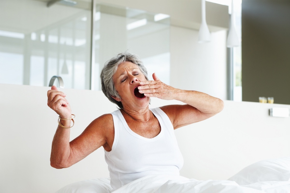 Senior woman sitting on bed and yawning with eyes closed