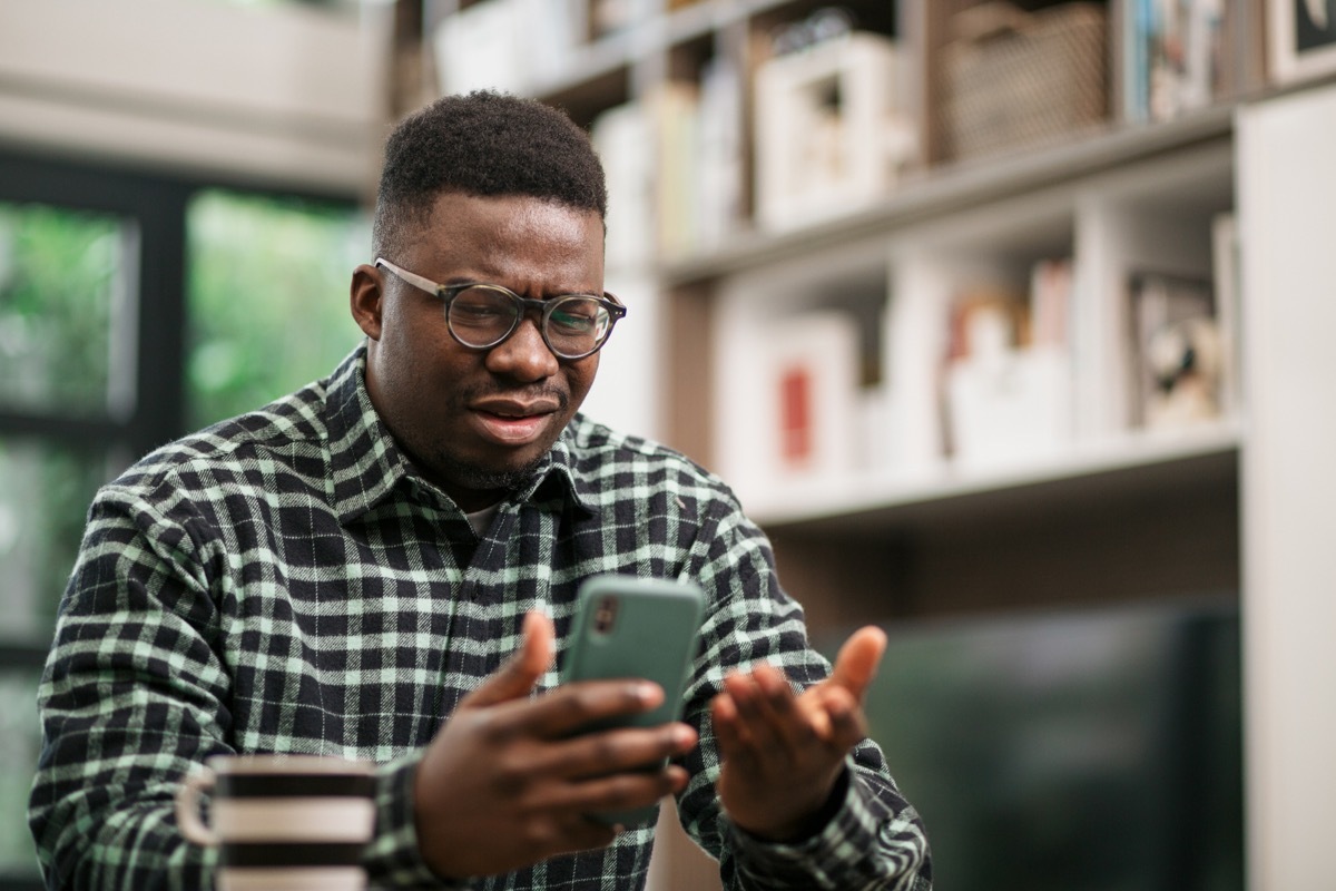 Annoyed young man sitting at home, reading some bad news on his smart phone using a mobile app and expressing his displeasure