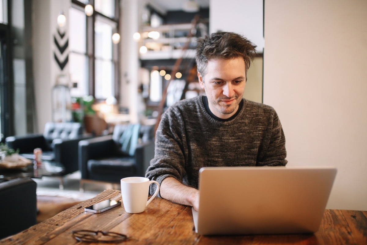 Young man sitting at the desk, doing an IT work or just casually browsing the web, checking e-mail, using social networks. He is having a cup of coffee, sitting in the living room of his Los Angeles loft apartment.