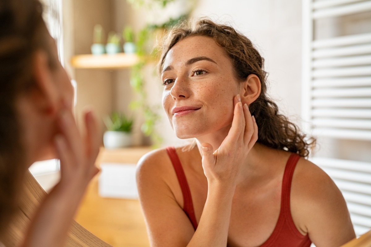 Beauty girl looking at mirror while touching her face and checking pimple, wrinkles and bags under the eyes, during morning beauty routine. Happy smiling beautiful young woman applying moisturizer.