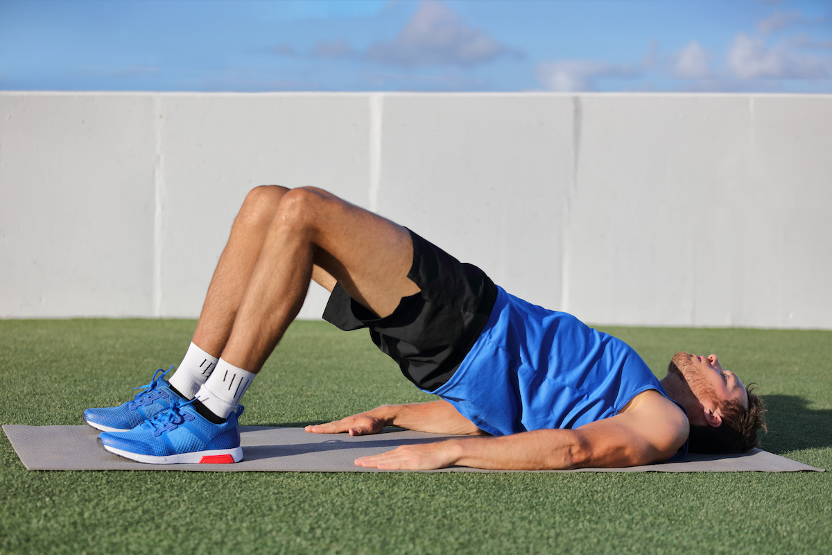 Fitness man doing bodyweight glute floor bridge pose yoga exercise. Fit athlete exercising glutes muscles with butt raise at summer outdoor gym instruction class on grass.