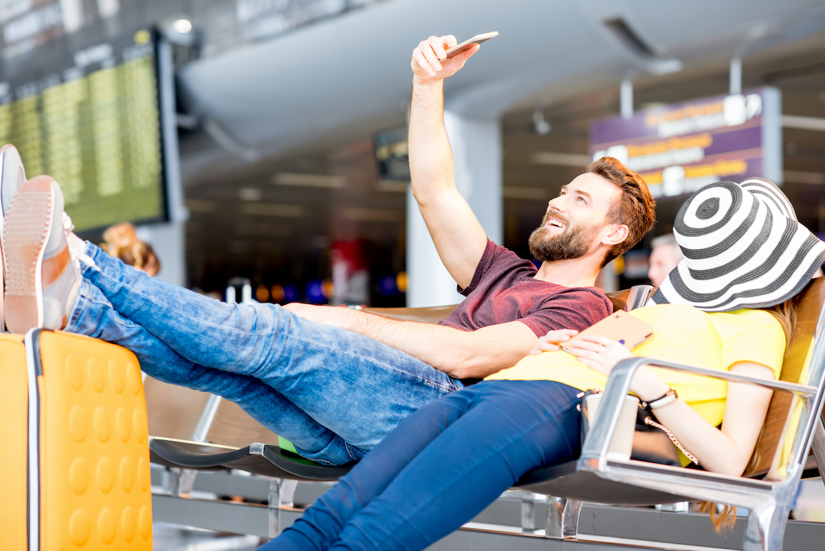 A young couple sits waiting at the airport; the man is smiling taking a selfie, while the woman has her hat over her face sleeping.