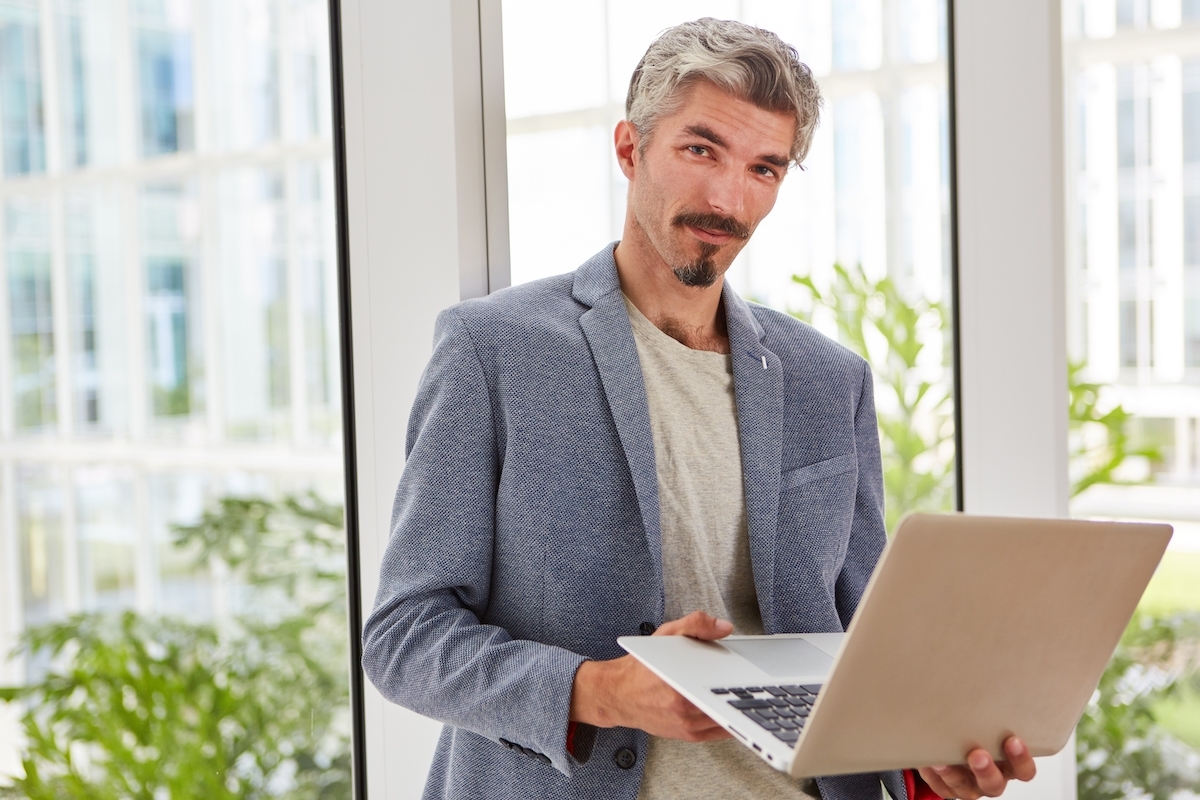 Portrait of confident businessman holding laptop against window. He has gray hair and is wearing a gray-blue blazer