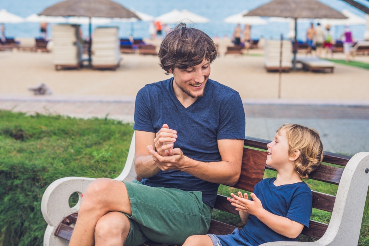 young white man in blue t-shirt and small boy using hand sanitizer on a bench near a beach