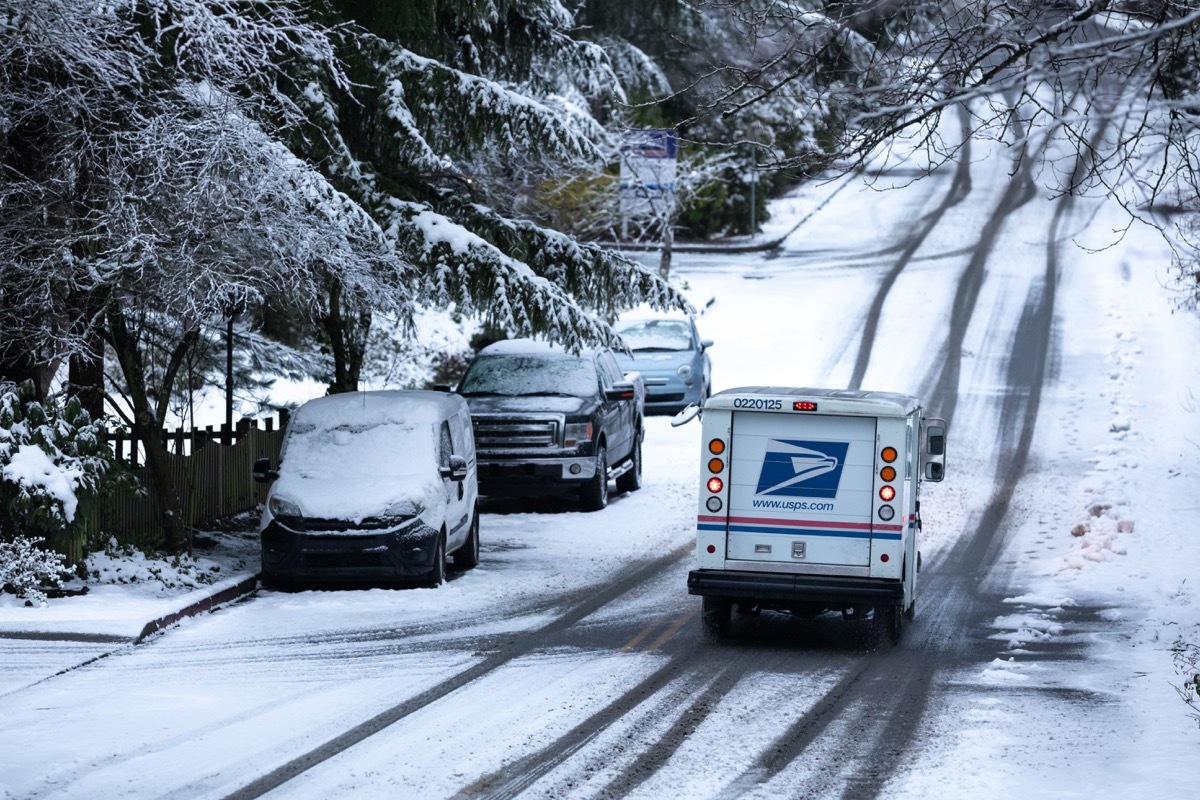 United States Postal Service (USPS) delivery truck driving down a snowy residential street during a winter storm, with space for text