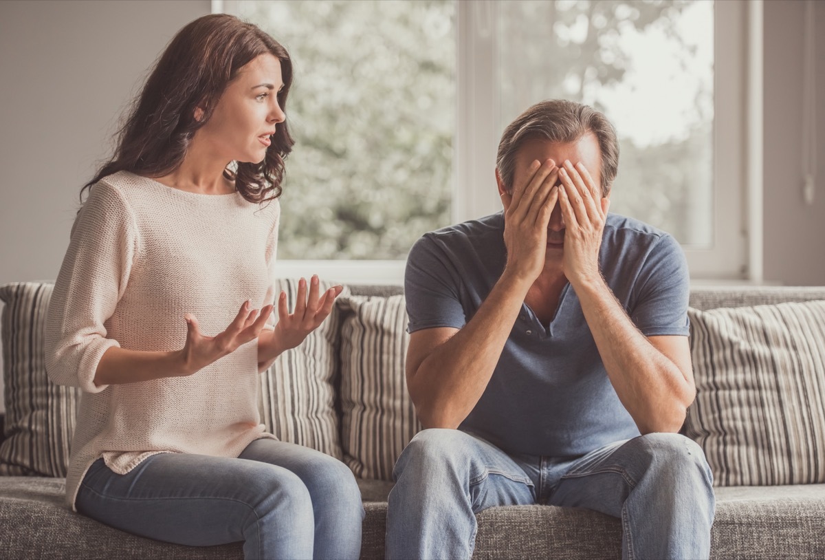 Couple of adults are quarreling while sitting on couch at home, man is covering his face