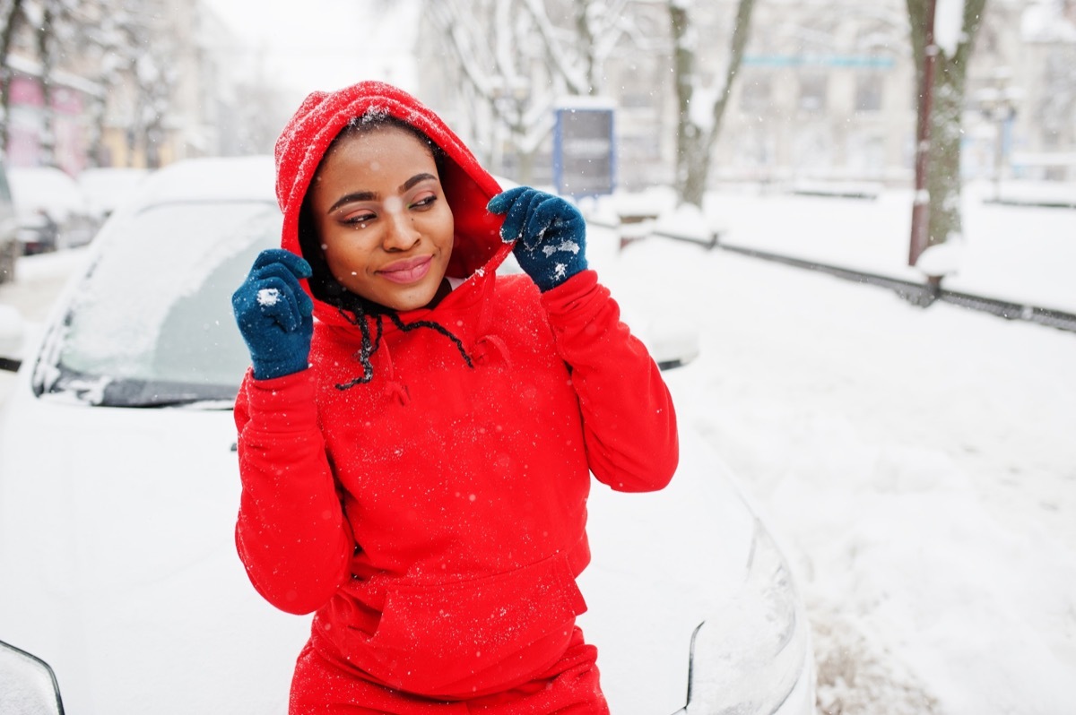 African american woman in red hoodie clean car from snow in winter day.