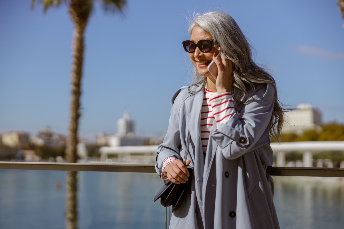 Mature woman walking next to water while talking on her phone. She's wearing a gray trench coat over a red-and-white striped shirt.