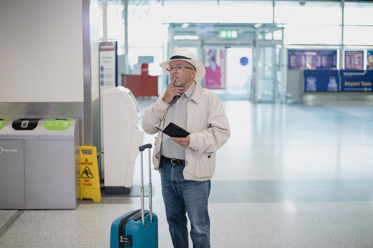 One senior man is standing in the airport terminal with his documentation, looking confusedly at the departure board.