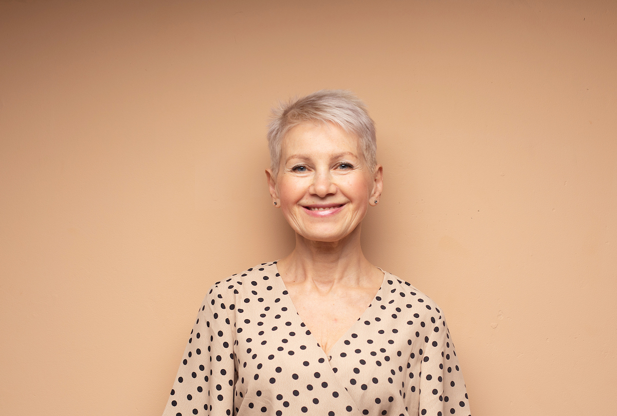 Mature smiling woman with a short pixie haircut wearing a beige dress with black polka dots against a beige background