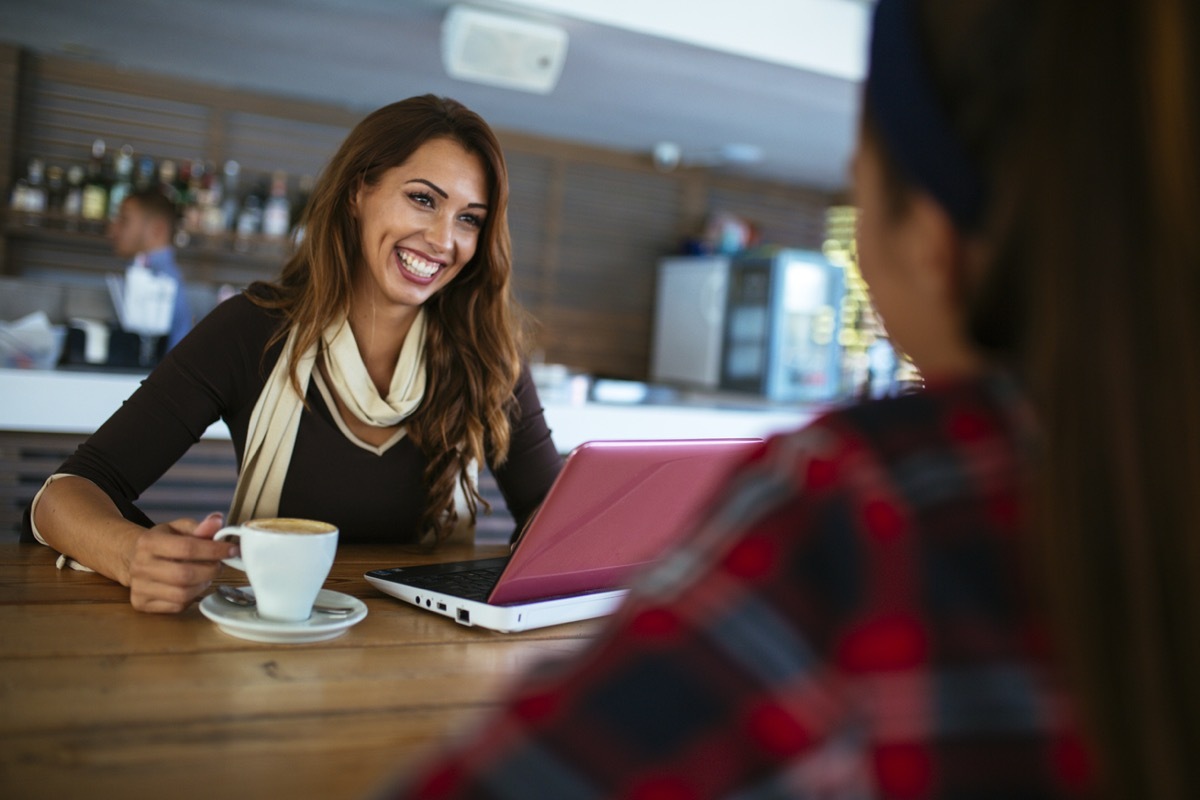 young woman smiling with her computer and coffee talking to another woman at a coffee shop