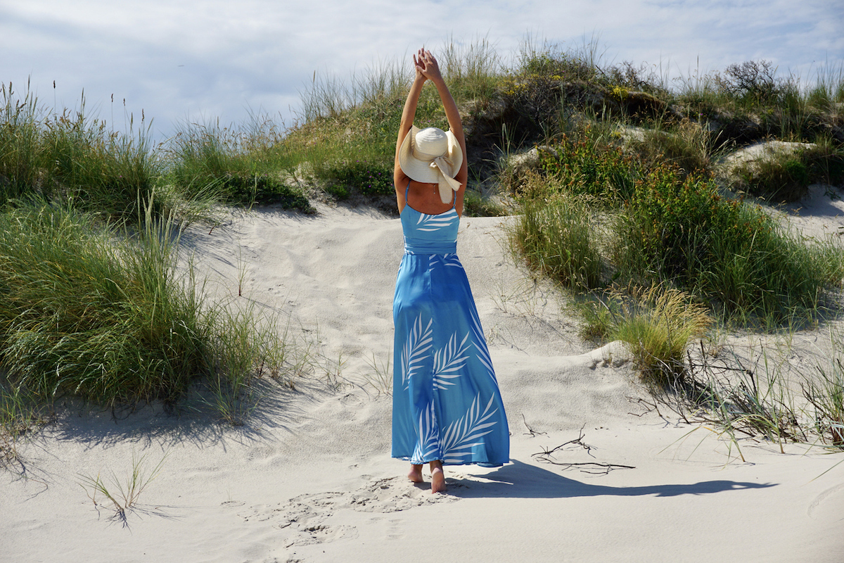 Rear view of a woman standing among sand dunes, wearing a long blue dress and a sun hat