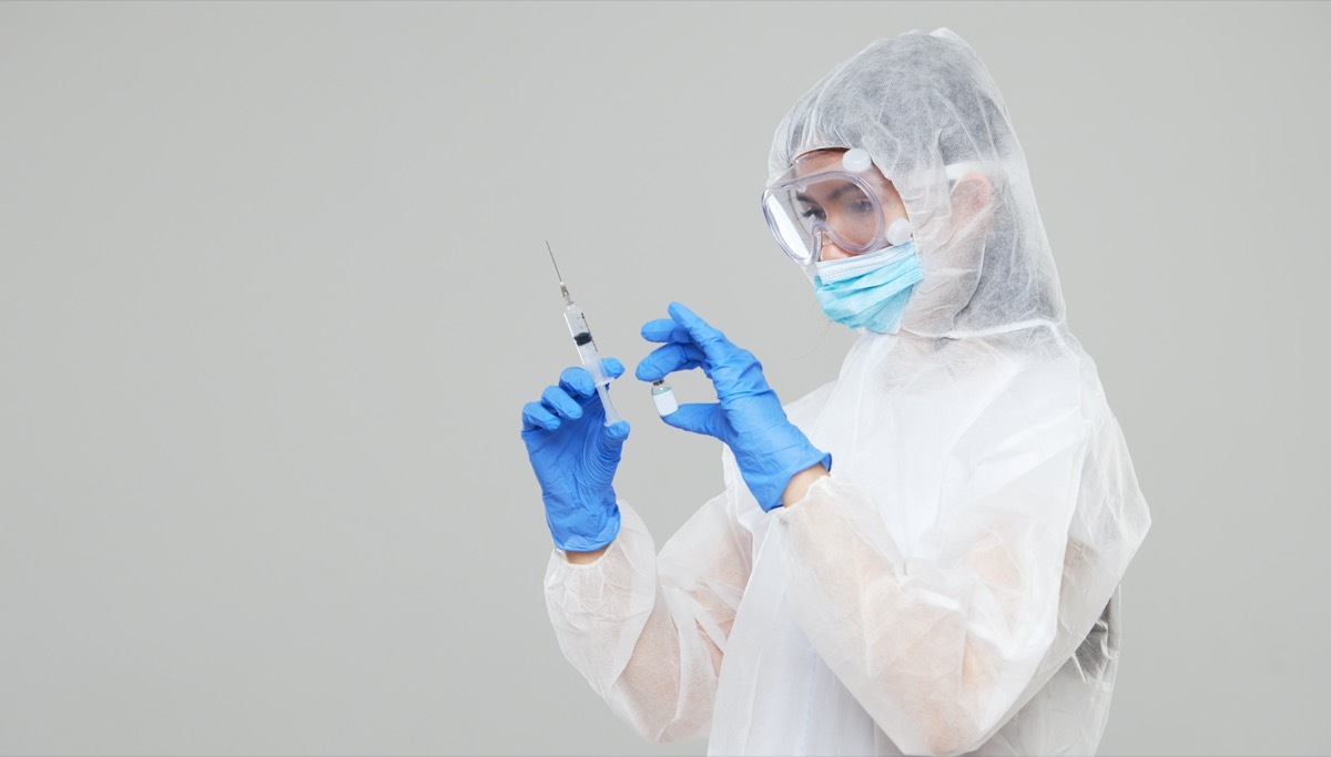 An asian woman in a protective suit and mask holds an injection syringe and vaccine