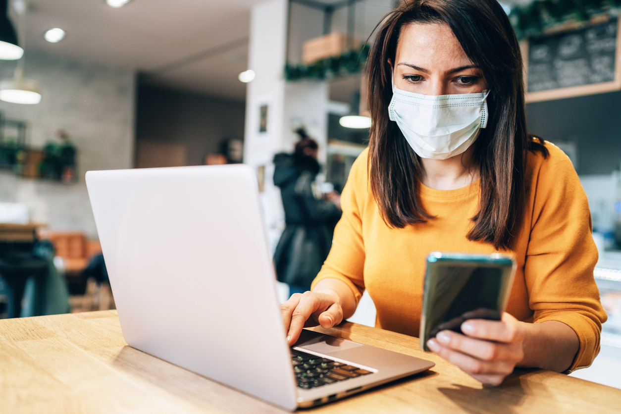 A young woman in a yellow sweater checks her smartphone while using her laptop and wearing a face mask