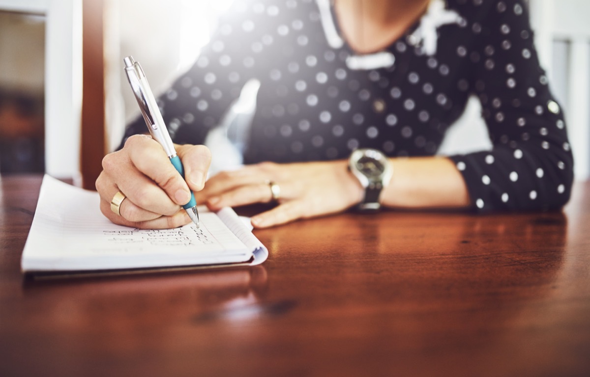 Shot of a unrecognizable woman writing in a book with a pen on a dinner table at home