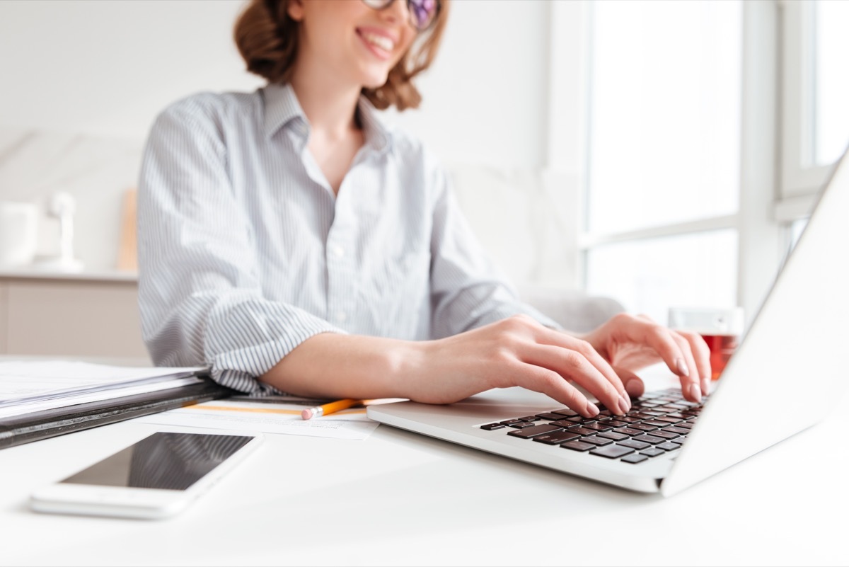 Cropped photo of pretty brunette woman typing email on laptop computer while sitting at home, selective focus on hand