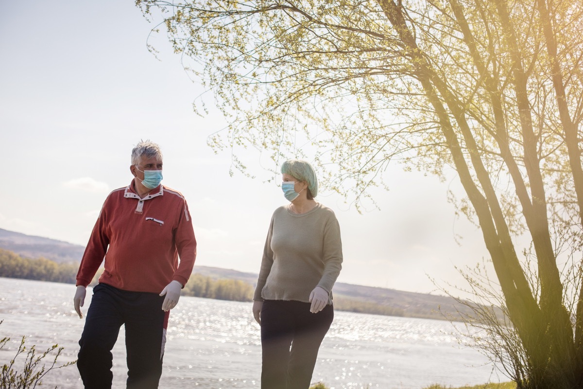 older white man and white woman walking by the water with face masks