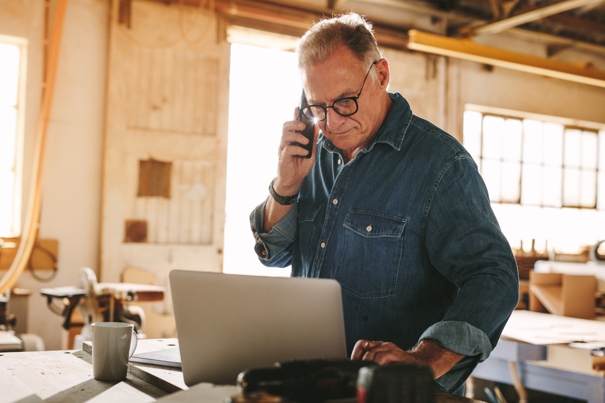 senior man talking on cell phone and using laptop on work table. Mature carpenter working on laptop and answering phone call in his carpentry workshop. (Senior man talking on cell phone and using laptop on work table.