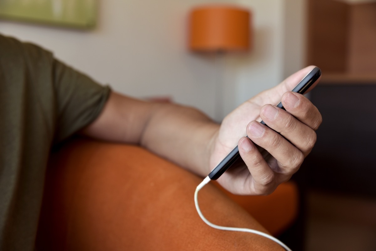 closeup of a young man using a smpartphone with a cable plugged into it, sitting in a comfortable sofa indoors