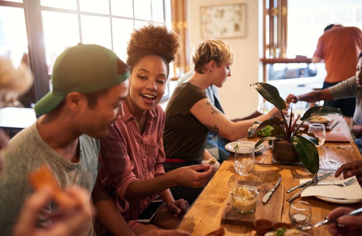 Diverse group of smiling young friends talking together over lunch and drinks at a table in a trendy bistro