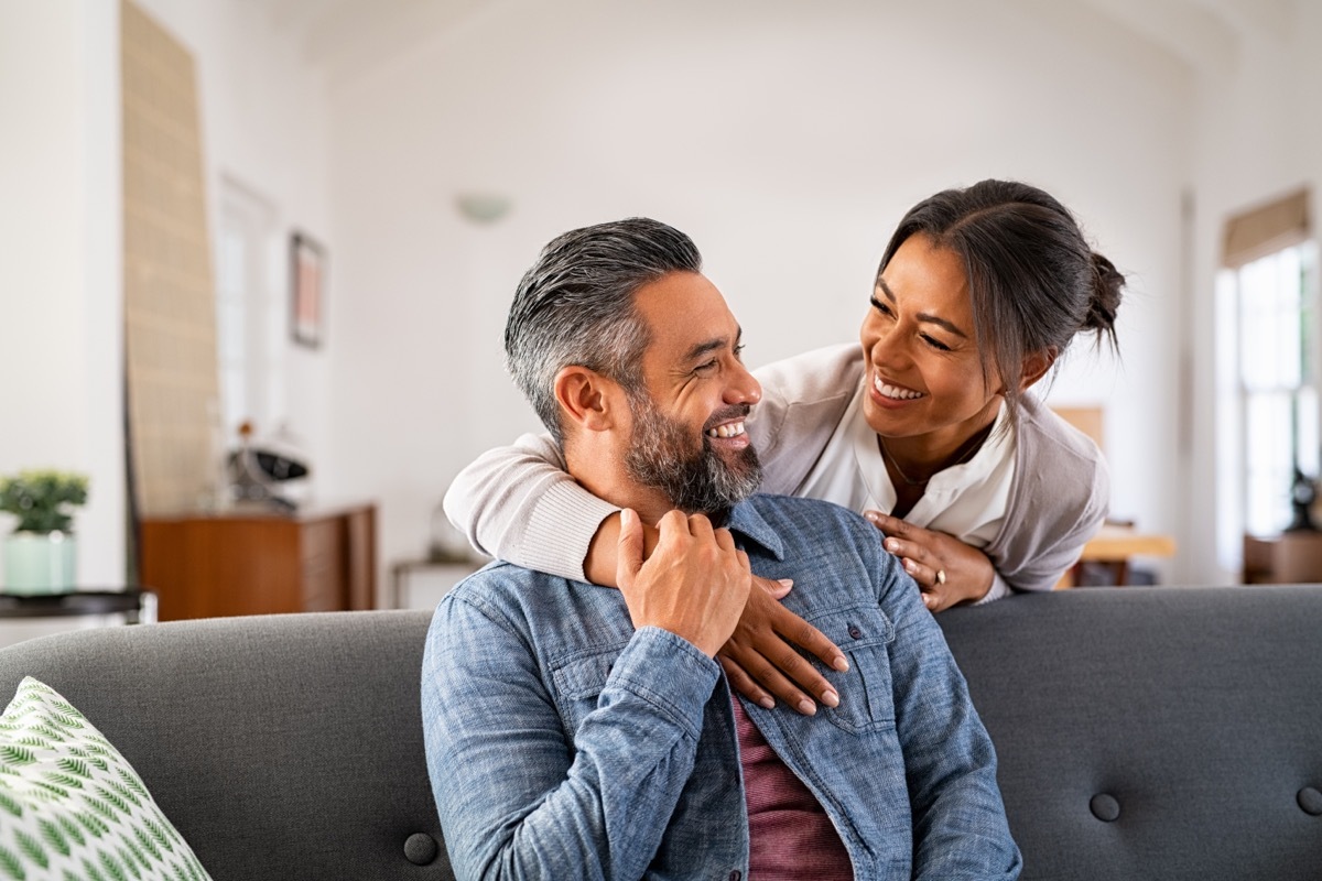 Smiling woman hugging her husband on the couch from behind in the living room. 