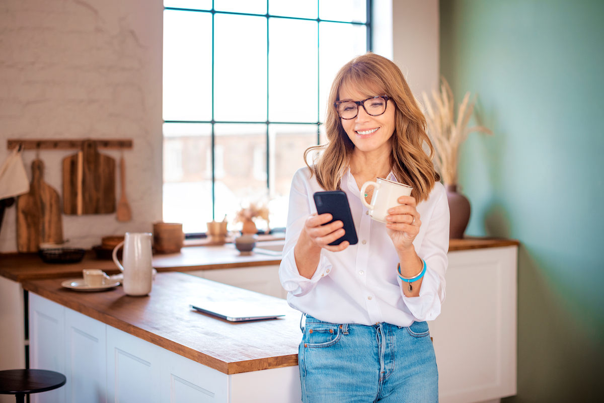 An attractive middle aged woman drinking her morning coffee and text messaging while relaxing at home in her kitchen.