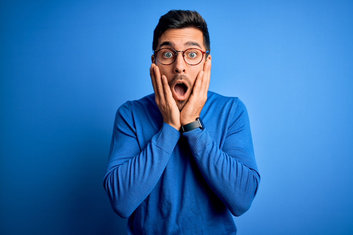 Young handsome man with beard wearing casual blue sweater and glasses over blue background afraid and shocked