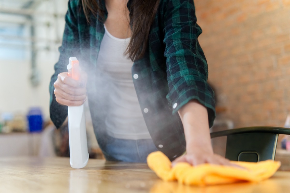 woman cleaning a house. She is wiping dust using a spray and a orange fabric while cleaning on the table