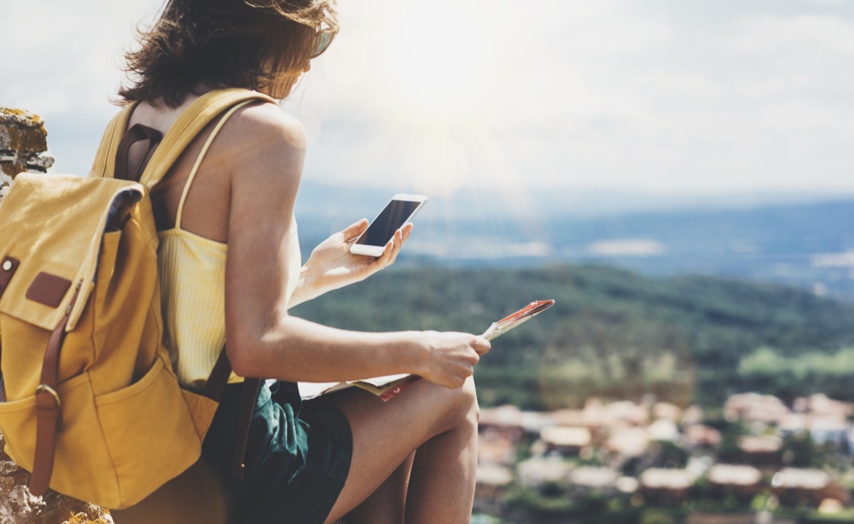 young female traveler with bright backpack looking at phone and map