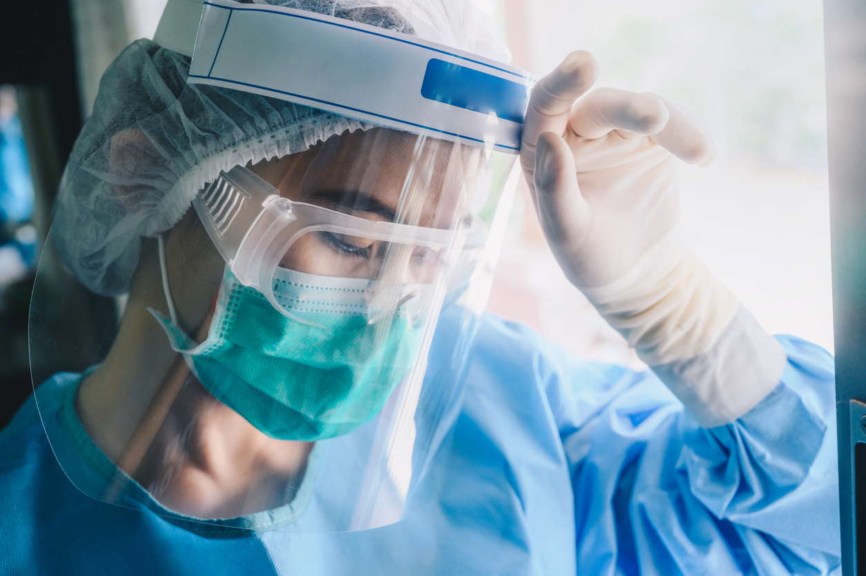 A young healthcare worker wearing a face shield and a face mask wears a worried expression while touching her forehead.