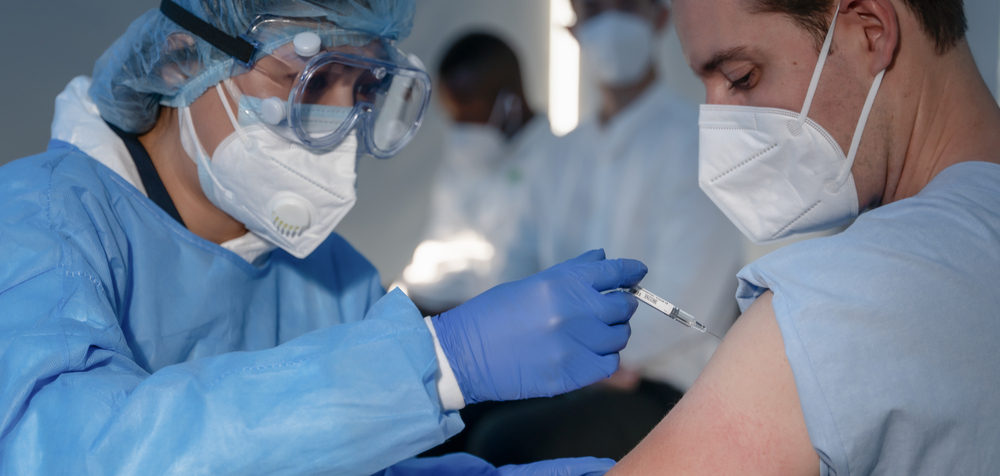 A health care worker wearing protective gear gives a young man wearing a face mask a COVID vaccination in his arm.