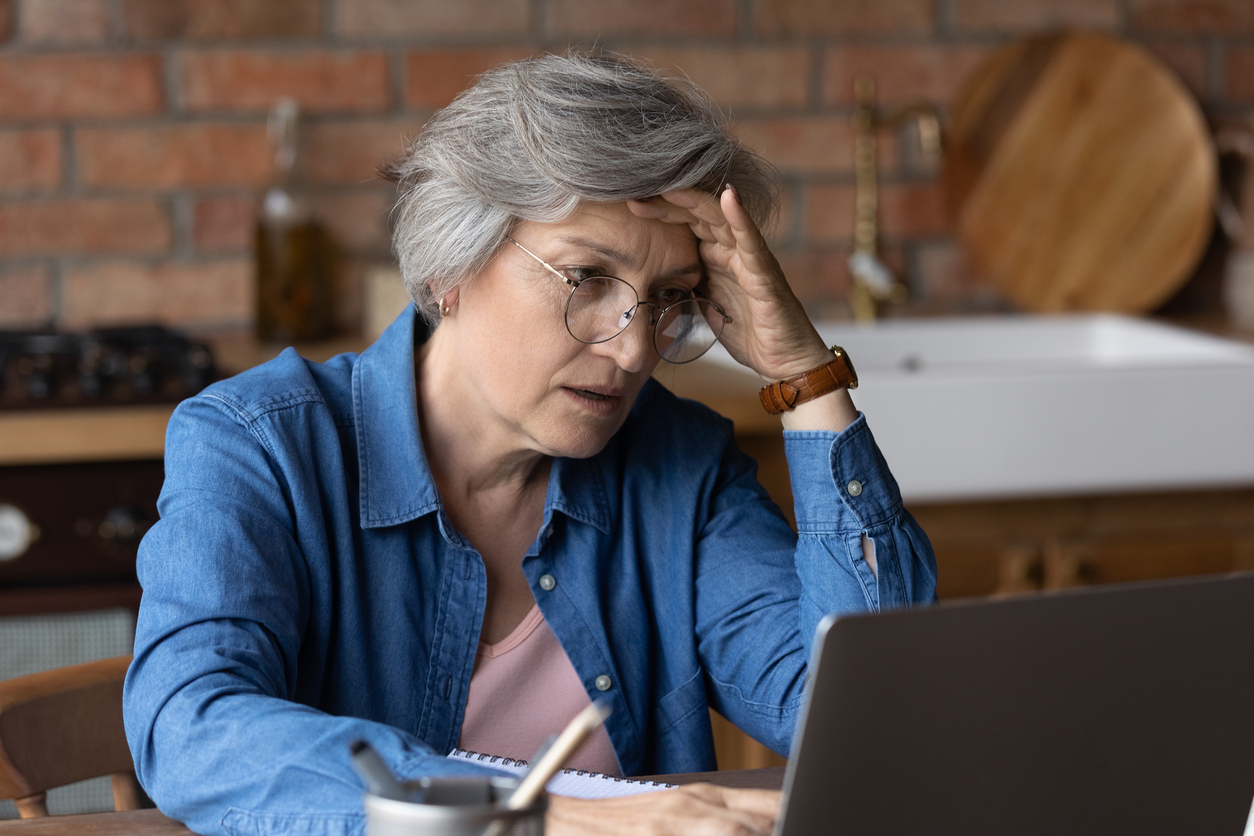 A senior woman sitting at her laptop in a kitchen with a distressed look on her face