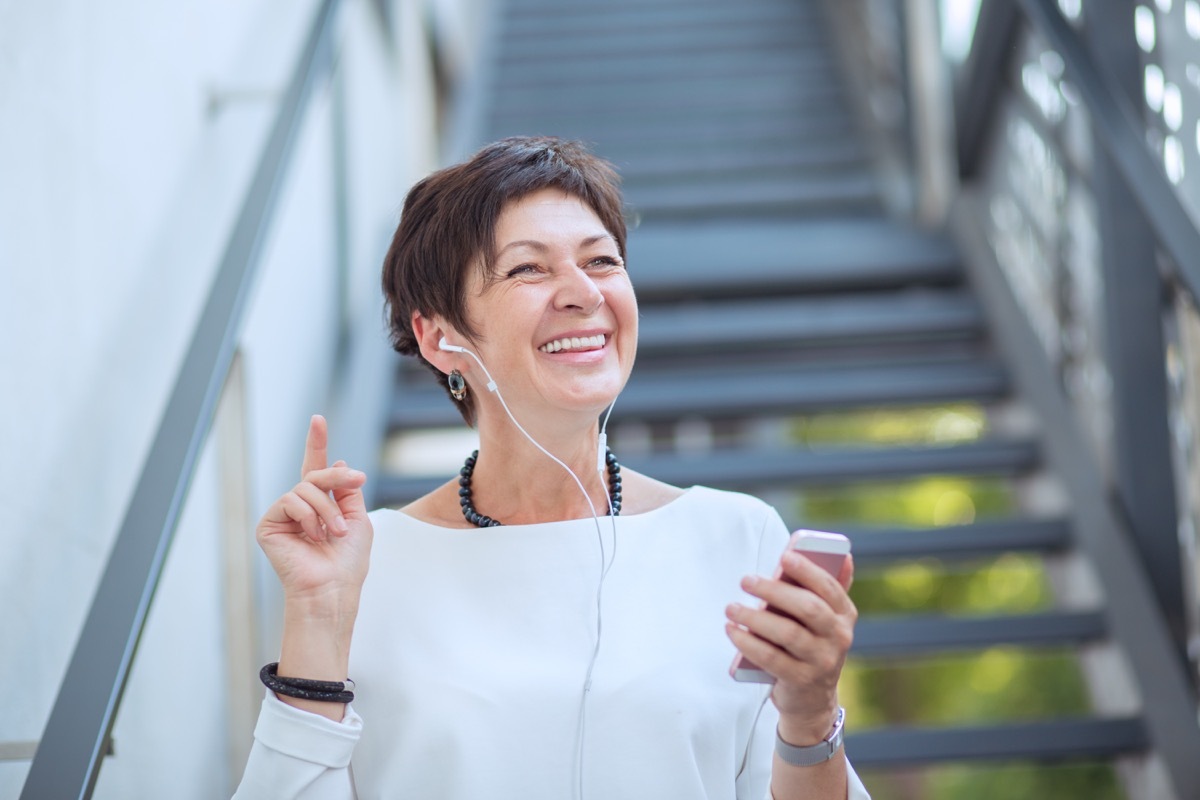 Content mature woman in trendy clothes listening to music with headphones and phone on street smiling away